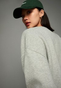 Grauer Pullover mit lockerem Schnitt und strukturiertem Stoff; kombiniert mit einer dunkelgrünen Kappe mit weißen Schriftzug. Das Model ist leicht zur Seite gedreht.