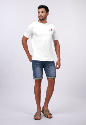 Λευκό βαμβακερό t-shirt με γραφικό ενός κόκκινου ήλιου, συνδυασμένο με ανοιχτό μπλε τζιν σορτς με διπλωμένες άκρες και καφέ σανδάλια.