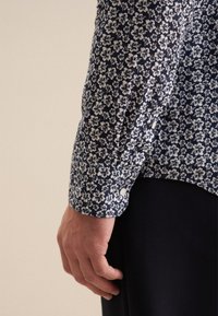 Camicia floreale blu navy con stampa bianca, caratterizzata da polsini con bottoni, tessuto morbido e vestibilità rilassata, che mostra un motivo floreale in scala ridotta.