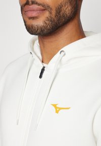 Biela mikina na zips s žltým logom na hrudi, obsahujúca šnúrky a kovové očká. Hladká tkanina.