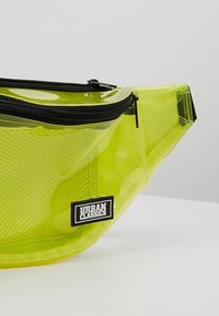Sacoche taille transparente néon vert avec fermeture éclair noire et étiquette "Urban Classics" à l'avant, présentée sur un fond uni.