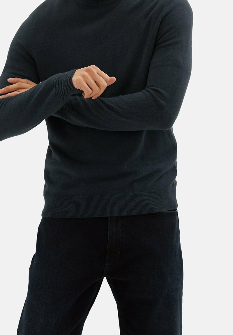 Personne portant un pull sombre à manches longues et un pantalon noir, les bras croisés devant un fond clair uni.
