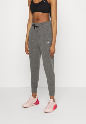 Person iført grå Puma joggers med snøre i taljen, sort sports-bh og lyserøde træningssko med røde detaljer, stående mod hvid baggrund.