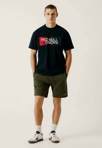 T-shirt preta com um logótipo "Red Bull Racing" em vermelho e branco. Calções verde-azeitona com um cós elástico. Ténis brancos com detalhes pretos.