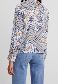 Bílá halenka s dlouhým rukávem a květinovým potiskem v modré, oranžové a hnědé barvě. Má výřez na zádech a vzor cikcak podél lemu.