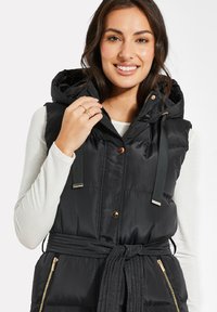 Schwarze gesteppten Weste mit Kapuze, mit tailliertem Gürtel, goldenen Hardware-Akzenten und Taschen mit Reißverschluss. Hergestellt aus glänzendem Stoff mit gestepptem Muster.