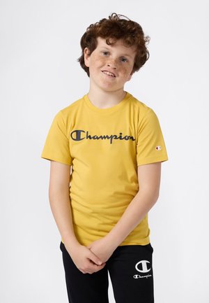 Geltona medvilnės marškinėliai trumpomis rankovėmis su juodu "Champion" logotipu krūtinėje; derinama su juodomis kelnėmis su baltu logotipu.