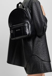 Czarny skórzany plecak o zaokrąglonym kształcie, z przednią kieszenią na zamek i wytłoczonym logo marki. Noszony na tle teksturowanej czarnej odzieży.