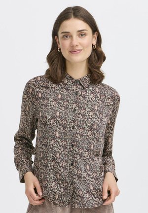 Frau mit schulterlangem braunem Haar lächelt und trägt ein Hemd mit einem mehrfarbigen floralen Paisleymuster auf hellem Hintergrund.