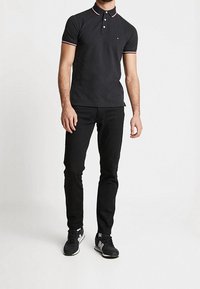 Man i svart kortärmad polo med röd och vit kant, svarta slim-fit byxor och svarta sportskor.