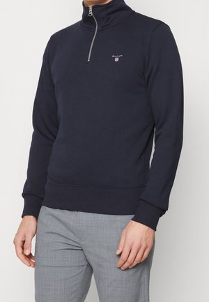 Mand iført en marineblå quarter-zip pullover med GANT-logo, kombineret med lysegrå mønstrede bukser, foran en ensfarvet baggrund.