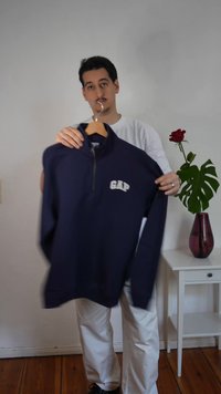 Moški v beli srajci drži mornarsko modri GAP pulover na lesenem obešalniku pred enobarvno steno ob stranski mizi z rdečo vrtnico v vazi.