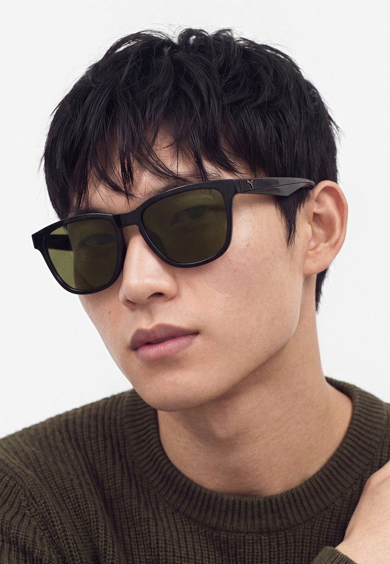 Giovane uomo con capelli neri corti che indossa occhiali da sole con montatura nera e un maglione di lana verde scuro, guarda leggermente di lato.