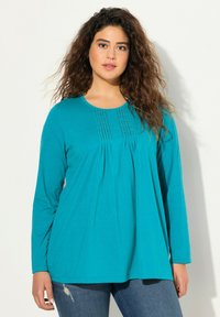 Teal langemouw top met geplooide details op de borst, gemaakt van zachte stof. Beschikt over een ronde halslijn en een losse, ontspannen pasvorm.