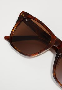 Ochelari de soare cu model țestoasă, cu o ramă îndrăzneață și unghiulară, lentile maro, puncte aurii de accent lângă balamale și un finisaj neted din plastic.