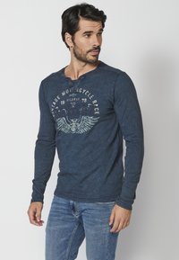 Dlouhý rukáv, modrá henley košile s kulatým výstřihem, texturovaná tkanina, knoflíkové detaily a grafika vintage motocyklu na přední straně.