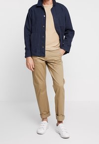 Uomo che indossa una giacca blu a bottoni, una maglietta beige, pantaloni khaki arrotolati e sneaker bianche, in piedi contro uno sfondo chiaro e uniforme.