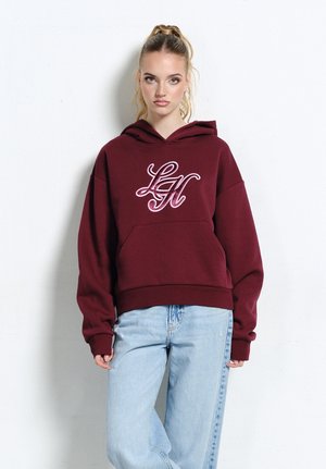 Młoda kobieta o blond włosach zebranych w wysoki kucyk, ubrana w bordową bluzę z kapturem z inicjałami "LH" i jasnoniebieskie jeansy, stoi na tle białej ściany.