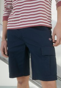 Osoba na sobě má námořnické cargo šortky s kapsami a pruhovanou košili s dlouhým rukávem, stojí s rukama podél těla.