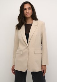 Beige bleiseri, jolla on rakenteellinen muotoilu, pistekuviointi, yksittäinen nappilukitus ja kaksi etutaskua. Päällä musta toppi.