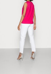 Fuchsia ärmelloses Oberteil kombiniert mit weißen, geraden Jeans, die fransige Säume haben. Als Schuhwerk werden silberne High-Heels getragen.