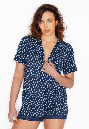 Femme aux cheveux ondulés portant une chemise courte à manches courtes bleu marine à boutons et un short assorti à pois blancs, tenant le col de la chemise.