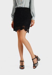 Gonna nera in pizzo con motivo floreale, orlo scallop, abbinata a sandali con tacco alto neri e argentati dotati di una tomaia testurizzata e cinturino alla caviglia.