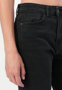 Svart denim jeans med høy midje, som viser en sidevinkel av en hånd nær lommen. Stoffet har en lett tekstur og synlige sømmer.