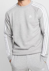 Man i ljusgrå Adidas träningsdräkt med vita ränder på ärmar och byxor, med händerna i fickorna, stående mot en enkel bakgrund.