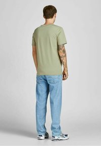 Vaaleanvihreä puuvillapaita, jossa on pyöreä kaula-aukko, yhdistettynä vaaleansinisiin farkkuihin. Malli esittelee takanäkymän, jossa ovat tatuoidut kädet.