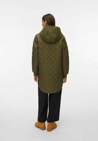 Podolg prešit olivno zelene jakne s kapuco, ki ima ukrivljen rob, rebraste manšete in gladko teksturo, kombiniran s črnimi širokimi hlačami in rjavimi škornji.