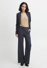 Marineblau gestreifter Blazer und weit geschnittene Hose kombiniert mit einem beigen Tanktop mit Spitzenbesatz. Das Material wirkt glatt und maßgeschneidert.