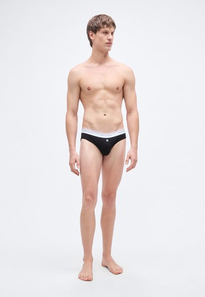 Slip noir avec une ceinture élastique blanche ornée d'un logo, fabriqué en tissu lisse, conçu pour une silhouette ajustée.