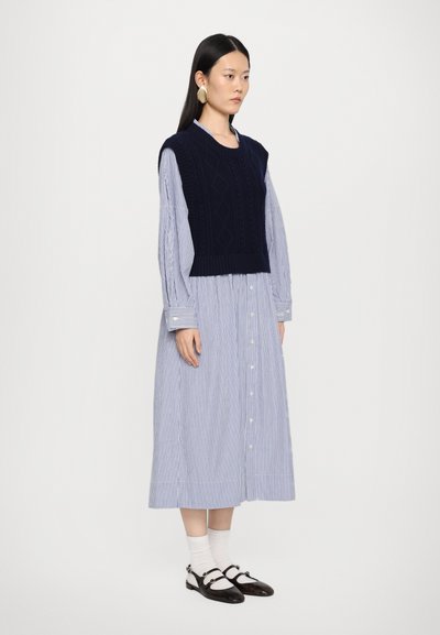 Dunkelblauer, kabelgenähter ärmelloser Pullover über einem blau-weißen gestreiften Langarm-Hemdblusenkleid, kombiniert mit schwarzen Schuhen und weißen Socken.