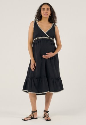 Femme enceinte portant une robe noire sans manches avec des finitions blanches, debout pieds nus sur un fond clair uni.