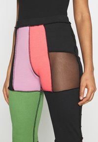 Femme portant des leggings color-block avec des panneaux en tissu vert, violet, corail et noir, et un panneau en mesh noir sur la partie supérieure de la cuisse.