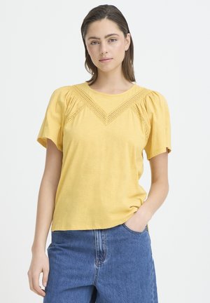 Junge Frau in gelbem Kurzarmtop mit Spitzenbesatz und blauer Jeans, steht mit einer Hand in der Tasche vor weißem Hintergrund.