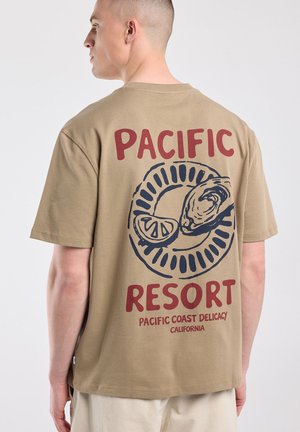 Man draagt een beige T-shirt met een print op de rug met oester, citroenschijfje en tekst "Pacific Resort Pacific Coast Delicacy California."