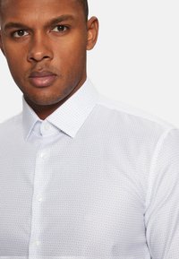 Camisa abotonada azul claro con un sutil patrón geométrico, que presenta un cuello clásico y botones blancos. El material tiene una textura suave.