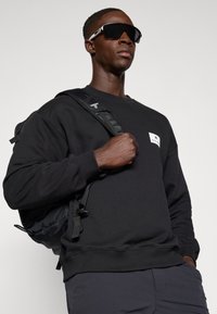 Felpa nera con scollatura rotonda, decorata con una patch logo bianca. Accompagnata da uno zaino nero e occhiali da sole neri oversize.