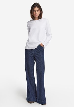 Witte lange mouwen overhemd gecombineerd met wijde donkerdenim jeans. De jeans hebben een afgewerkte zoom met een lichte versleten rand onderaan.