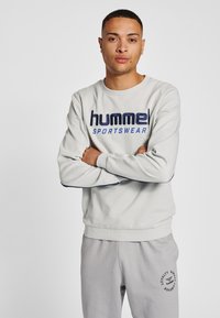 Jasnoszara bluza z ciemnoniebieskim napisem "hummel SPORTSWEAR", z prążkowanymi mankietami i krawędzią, noszona z dopasowanymi jasnoszarymi dresami z detalem logo.