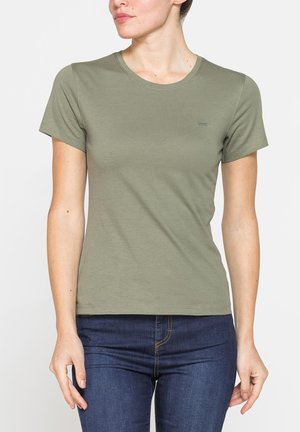 Donna con maglietta verde oliva a maniche corte semplice e jeans blu scuro, in piedi davanti a uno sfondo chiaro e uniforme.
