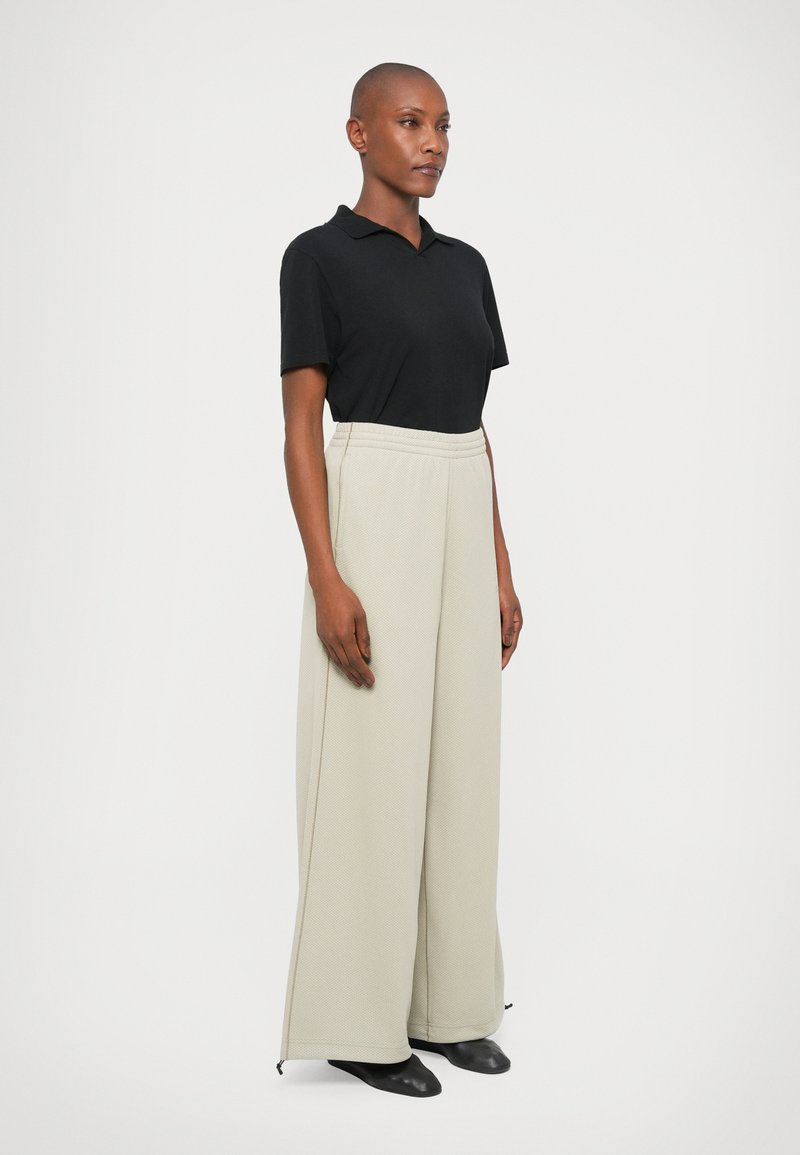 Chemise polo noire avec col, associée à un pantalon léger beige lâche et texturé. Le pantalon possède une taille à cordon et des zips au bas.