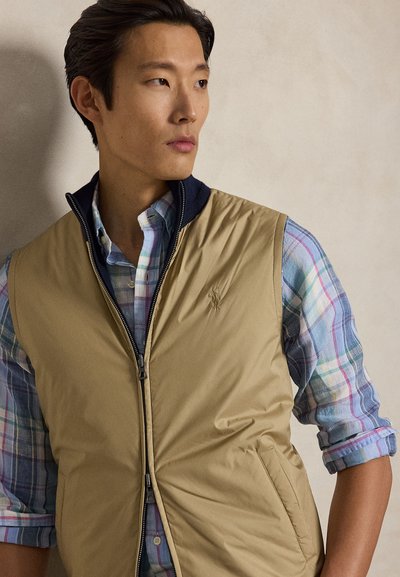 Jauns vīrietis valkā bēša zipša vestīti virs pasteļtoņu pleda krekla, skatoties uz sāniem pret neitrālu fonu.