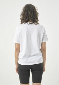 Weißes Baumwoll-T-Shirt mit kurzen Ärmeln, schlichter Rücken, lockere Passform. Getragen mit engen schwarzen Shorts. Neutraler Hintergrund.