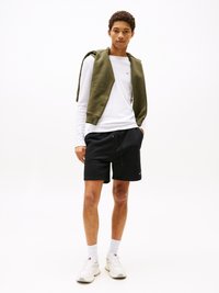 Czarne spodenki i biała koszula z długim rękawem, na to nałożona oliwkowa kamizelka bez rękawów. Obuwie: białe sneakersy. Czysty, casualowy wygląd.