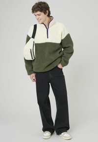 Pull en polaire avec une partie supérieure crème et une partie inférieure vert olive, agrémenté d'une fermeture éclair violette et d'une coupe décontractée. Associé à un jean noir et des baskets.