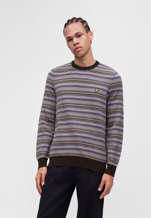 Jeune homme aux cheveux tressés portant un pull rayé multicolore avec un petit logo jaune sur la poitrine et un pantalon noir.