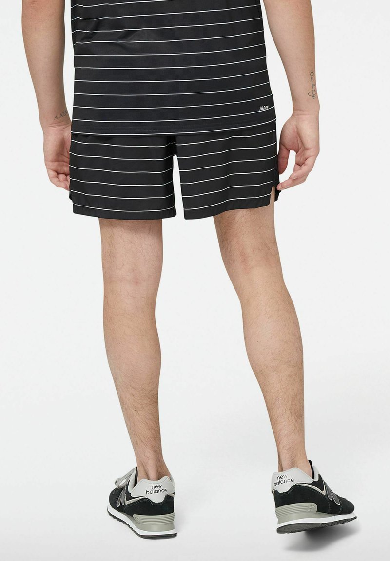 Schwarze Shorts mit weißen horizontalen Streifen, kombiniert mit schwarzen New Balance Sneakers. Sichtbare Beinbehaarung und ein Tattoo am linken Arm.